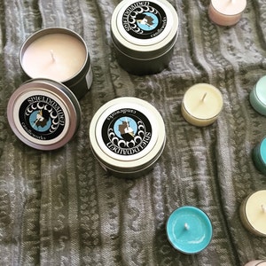 May include: Two round silver metal candle tins with lids featuring a black circular label with the text SHIELDMAIDEN candleworks and a profile silhouette. One open tin reveals a pale pink wax candle. Several small tea light candles in various colors.