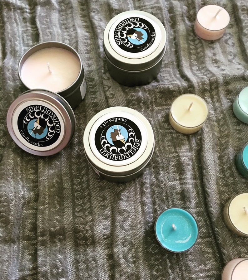 May include: Two round silver metal candle tins with lids featuring a black circular label with the text SHIELDMAIDEN candleworks and a profile silhouette. One open tin reveals a pale pink wax candle. Several small tea light candles in various colors.
