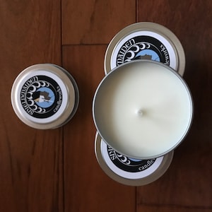 May include: Several small, round, silver-colored candle tins are arranged on a wooden surface. One tin is open, revealing a creamy white candle with a single wick. The lids feature a circular label with the text "Spiced Maiden Candleworks".