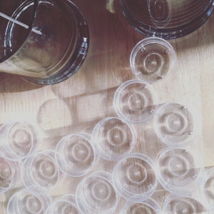 May include: A collection of clear, circular plastic candle containers and a single glass candle vessel with a centered wick. The plastic containers are arranged in a scattered pattern, featuring a recessed base and a small central circular indentation.
