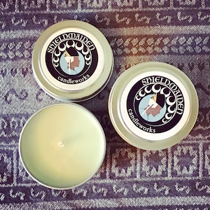 May include: Two round silver metal candle tins with lids and one open candle tin containing pale green wax and a centered wick. The lids feature a circular logo with the text SHIELDMAIDEN candleworks and an illustration of a profile silhouette.