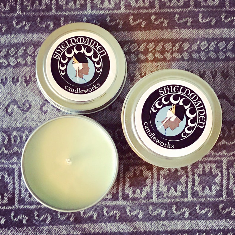 May include: Two round silver metal candle tins with lids and one open candle tin containing pale green wax and a centered wick. The lids feature a circular logo with the text SHIELDMAIDEN candleworks and an illustration of a profile silhouette.