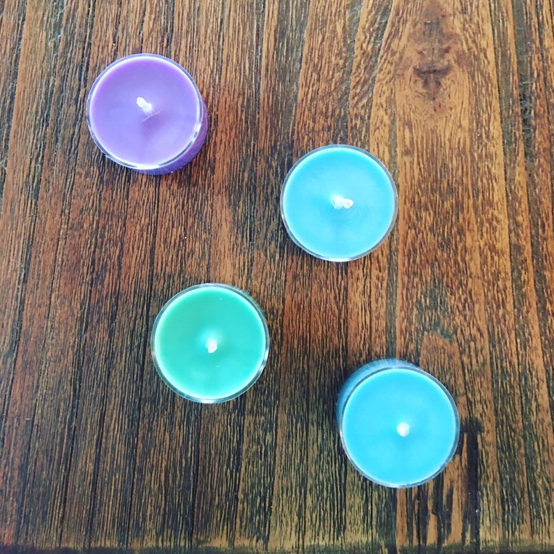 May include: Four round tea light candles in clear plastic cups, arranged in a scattered pattern. The candles feature smooth wax surfaces in shades of purple, light blue, and green, each with a centered white cotton wick ready for lighting.