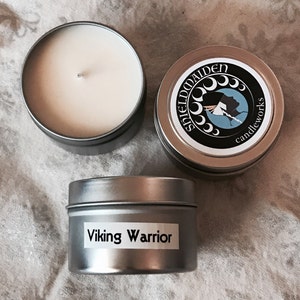 May include: Three silver-colored candle tins on a patterned surface. One tin is open, revealing a white candle. The other two tins have labels. One label reads "Shieldmaiden candleworks" and the other reads "Viking Warrior".