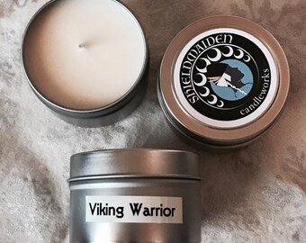 May include: Three silver-colored candle tins on a patterned surface. One tin is open, revealing a white candle. The other two tins have labels. One label reads "Shieldmaiden candleworks" and the other reads "Viking Warrior".