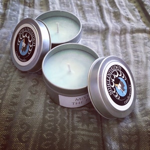May include: Two round silver metal candle tins with light blue wax and white wicks. Each lid features a circular black label with the text SHIELDMAIDEN candleworks and a graphic of a profile silhouette. One tin has a partial white side label.