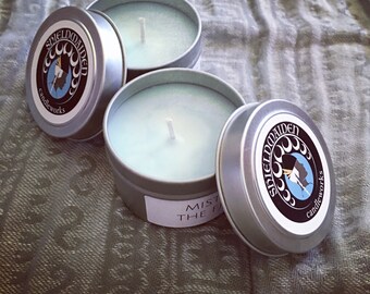 May include: Two round silver metal candle tins with light blue wax and white wicks. Each lid features a circular black label with the text SHIELDMAIDEN candleworks and a graphic of a profile silhouette. One tin has a partial white side label.