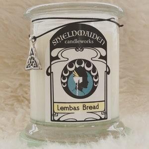 May include: A clear glass jar candle with a black label that reads "Shieldmaiden candleworks" and "Lembas Bread". A silver charm hangs from the jar. The candle is white and has a black string tied around the top.