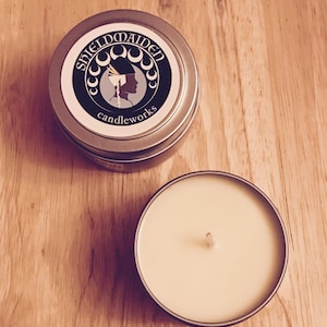 May include: Two silver candle tins on a wooden surface. One tin is open, revealing a cream-colored candle with a wick. The closed tin has a label with the text "SHIELDMAIDEN candleworks" and a silhouette of a woman.
