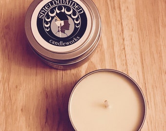 May include: Two silver candle tins on a wooden surface. One tin is open, revealing a cream-colored candle with a wick. The closed tin has a label with the text "SHIELDMAIDEN candleworks" and a silhouette of a woman.