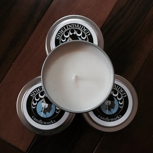 May include: Four silver candle tins with white wax and a wick. The lids feature a black and white design with the text "SHIELDMAIDEN candleworks" and a silhouette. The candles are arranged on a wooden surface.