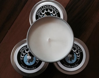 May include: Four silver candle tins with white wax and a wick. The lids feature a black and white design with the text "SHIELDMAIDEN candleworks" and a silhouette. The candles are arranged on a wooden surface.