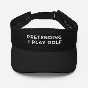 Könnte beinhalten: Schwarzes Visier mit dem gestickten Text "PRETENDING I PLAY GOLF" in Weiß. Das Visier hat einen verstellbaren Riemen und einen gebogenen Schirm. Das Flexfit-Markenlogo ist sichtbar.