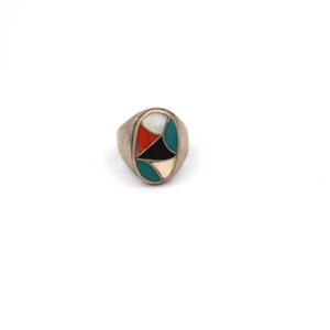 May include: Silver-toned ring with an oval face featuring a geometric design. The design incorporates red, black, white, and teal-colored inlays. The ring has a polished finish and a classic, unisex style.
