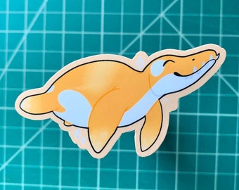 May include: A sticker featuring a cartoon plesiosaur in shades of orange and light blue. The plesiosaur has a smiling expression and is outlined in black. The sticker is on a green cutting mat.