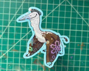 May include: A sticker featuring a cartoon pterodactyl with a light blue outline. The pterodactyl has a white body, brown wings with snowflake designs, and a black hat. The background is a green cutting mat.