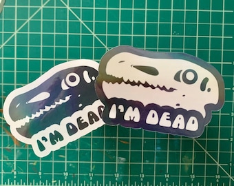 May include: Two stickers featuring a dinosaur skull design with the text "I'M DEAD". The stickers have a holographic, iridescent finish, with colors shifting from blue to purple. The design is outlined in white.