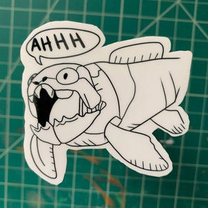 May include: A white sticker featuring a cartoon Coelacanth fish with the word "AHHH" in a speech bubble. The fish has large teeth and is outlined in black. The sticker is on a green cutting mat.