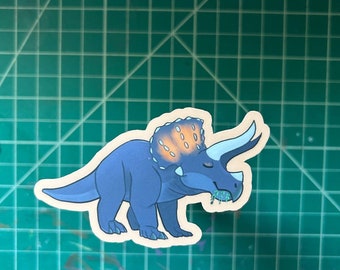 May include: A die-cut sticker featuring a blue triceratops dinosaur with a light blue horn and a frill with orange accents. The dinosaur is eating a small green insect. The sticker is on a green cutting mat.