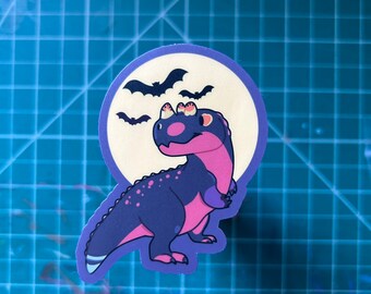 May include: A sticker featuring a cartoon dinosaur with a purple and pink color scheme, set against a cream-colored moon and a purple border. Above the dinosaur are several black bat silhouettes, creating a Halloween theme.