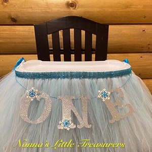May include: A light blue tulle high chair skirt with a silver glitter "ONE" banner and blue snowflake embellishments. The skirt is decorated with a blue and silver sparkly trim. The text "Nonna's Little Treasures" is printed on the skirt.