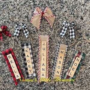May include: Six personalized Christmas ornaments with names spelled out using letter tiles. Each ornament is made with a wooden plaque, burlap ribbon, and a plaid bow. The names are: Bella, Kacey, Juliana, Mason, Noel.