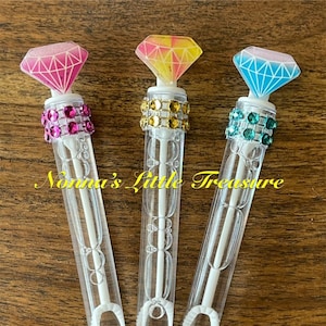 May include: Three clear plastic bubble wands with diamond-shaped tops. The tops are pink, yellow, and blue with glitter accents. Each wand has a ring of colorful rhinestones around the middle.