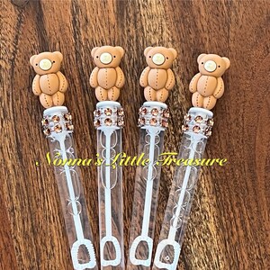 May include: Four clear plastic bubble wands with brown teddy bear toppers and a white wand. Each wand has a clear plastic container filled with bubble solution and a white wand with a loop at the end. The wands are decorated with brown and gold glitter.