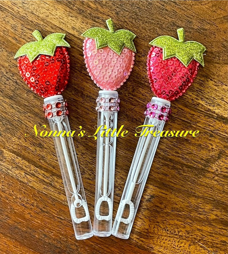 May include: Three clear plastic bubble wands with a strawberry-shaped topper. The toppers are made of red, pink, and light pink sequins with green glitter leaves. Each wand has a clear plastic tube with a small loop at the bottom and a clear plastic handle.