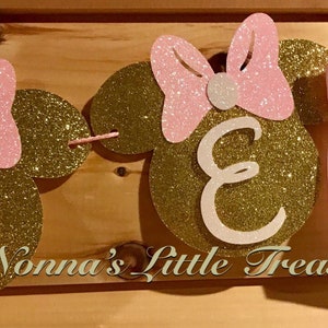 May include: Gold glitter Minnie Mouse themed banner with pink bows and the letter "N" in white. The banner says "Nonnas Little Treasure".