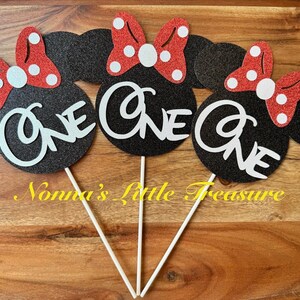 May include: Three black glittery mouse-shaped cake toppers, each featuring a red glittery bow with white polka dots and the word One in white script lettering. Each topper is attached to a thin white stick for placement in a cake or dessert.
