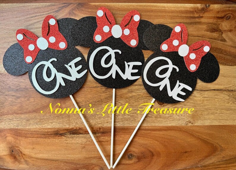 May include: Three black glittery mouse-shaped cake toppers, each featuring a red glittery bow with white polka dots and the word One in white script lettering. Each topper is attached to a thin white stick for placement in a cake or dessert.