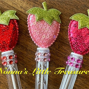 May include: Three clear plastic bubble wands with a strawberry-shaped topper. Each strawberry is covered in sequins and has a green glitter leaf. The wands are decorated with a band of rhinestones.