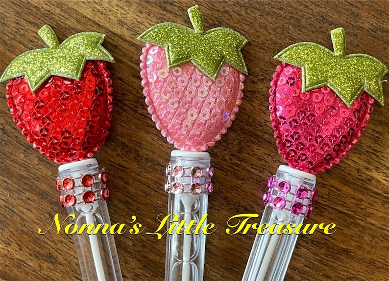May include: Three clear plastic bubble wands with a strawberry-shaped topper. Each strawberry is covered in sequins and has a green glitter leaf. The wands are decorated with a band of rhinestones.