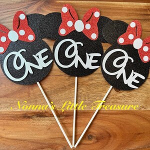 May include: Three black glittery mouse-shaped cake toppers, each featuring a red glittery bow with white polka dots and the word ONE in white script lettering. Each topper is attached to a thin white stick for placement in a cake or dessert.