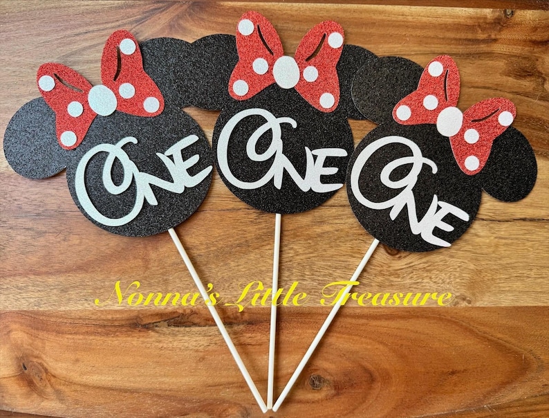 May include: Three black glittery mouse-shaped cake toppers, each featuring a red glittery bow with white polka dots and the word ONE in white script lettering. Each topper is attached to a thin white stick for placement in a cake or dessert.