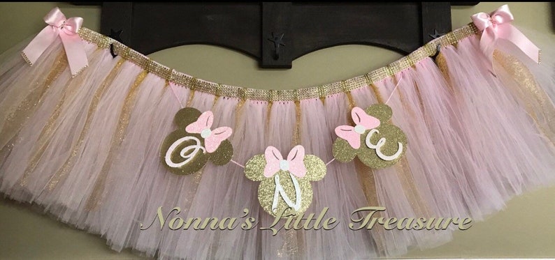 May include: Pink and gold tulle tutu with a banner that says "One" in white letters with pink and gold Minnie Mouse ears.