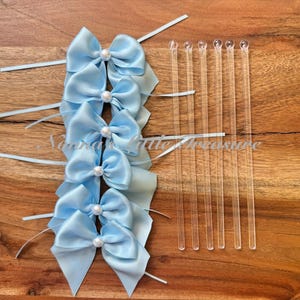 May include: A collection of light blue satin bows with pearl accents and clear plastic stir sticks. The bows are arranged vertically on a wooden surface. The bows have long, thin ribbons attached.