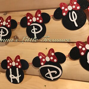 May include: A banner featuring black Minnie Mouse head cutouts with red and white polka dot bows. Each head has a white letter or symbol. The banner is strung on red string and is displayed against a wooden background.