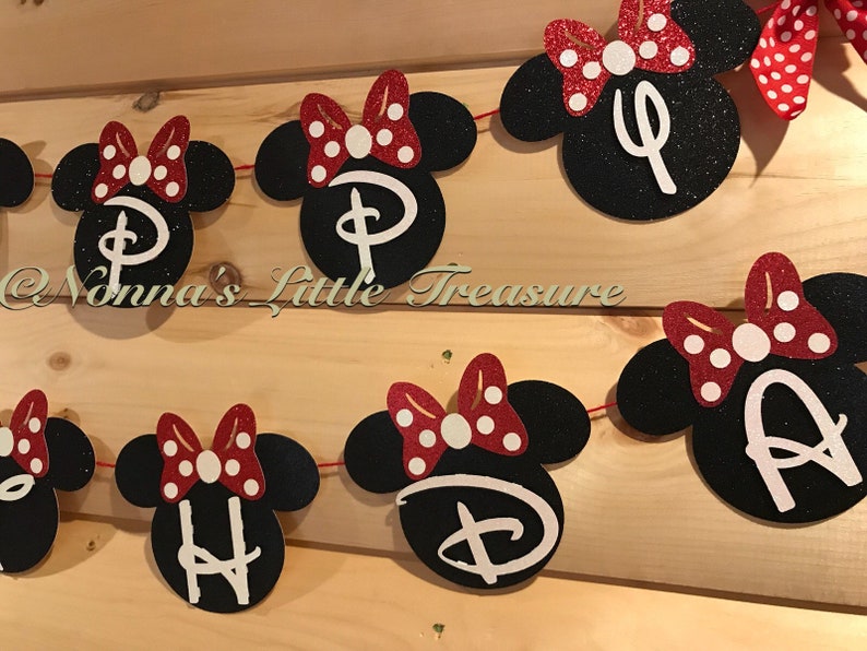 May include: A banner featuring black Minnie Mouse head cutouts with red and white polka dot bows. Each head has a white letter or symbol. The banner is strung on red string and is displayed against a wooden background.