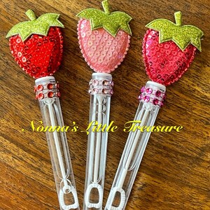 May include: Three clear plastic bubble wands with a red, pink, and hot pink sequin strawberry on top. The wands have a clear plastic handle and a small loop at the bottom. The strawberries have green glitter leaves.