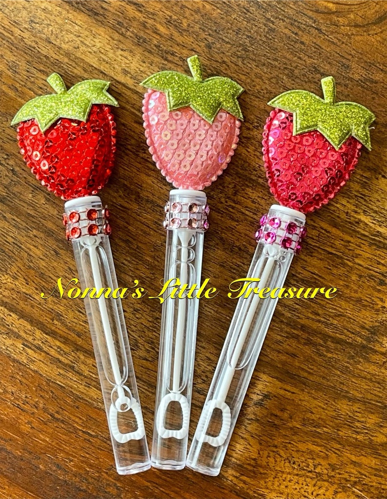 May include: Three clear plastic bubble wands with a red, pink, and hot pink sequin strawberry on top. The wands have a clear plastic handle and a small loop at the bottom. The strawberries have green glitter leaves.