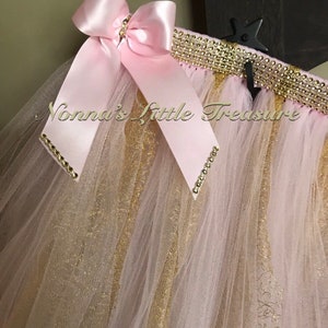 May include: A pink tulle skirt with gold glitter and a pink satin bow. The skirt has a gold and rhinestone trim.