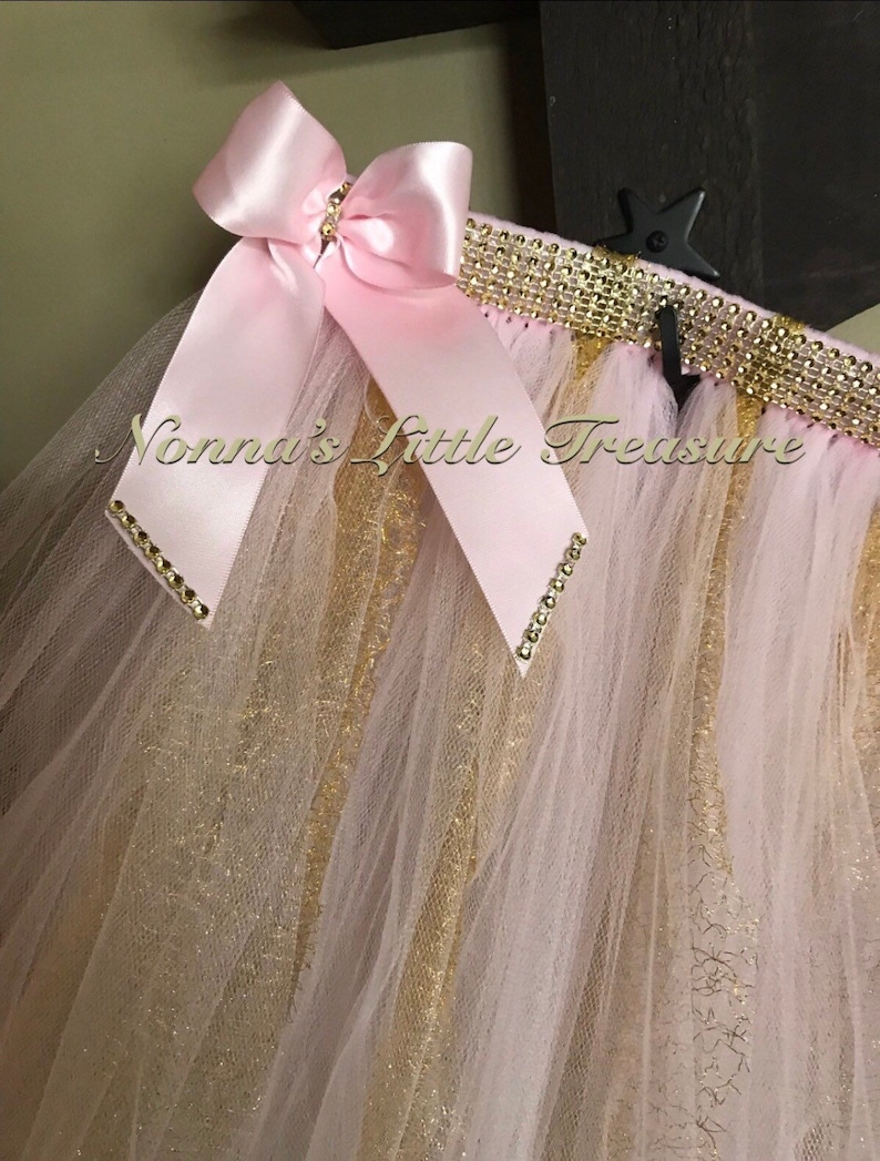 May include: A pink tulle skirt with gold glitter and a pink satin bow. The skirt has a gold and rhinestone trim.