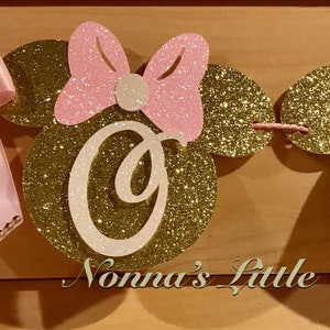 May include: A pink and gold glitter banner with the letters "ONE" in white. Each letter is on a gold glitter mouse head with pink glitter ears and a pink bow. The banner is decorated with a pink satin bow.