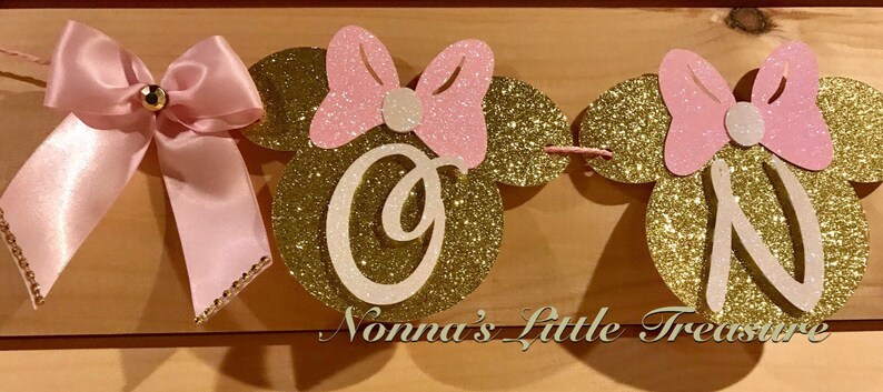 May include: A pink and gold glitter banner with the letters "ONE" in white. Each letter is on a gold glitter mouse head with pink glitter ears and a pink bow. The banner is decorated with a pink satin bow.