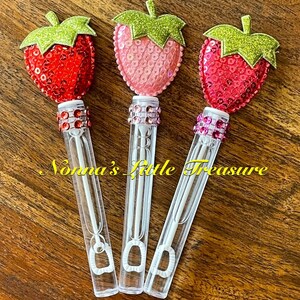 May include: Three bubble wands with strawberry-shaped tops. The tops are made of red, pink, and fuchsia sequins with green glitter leaves. The wands are clear plastic with a white wand inside.
