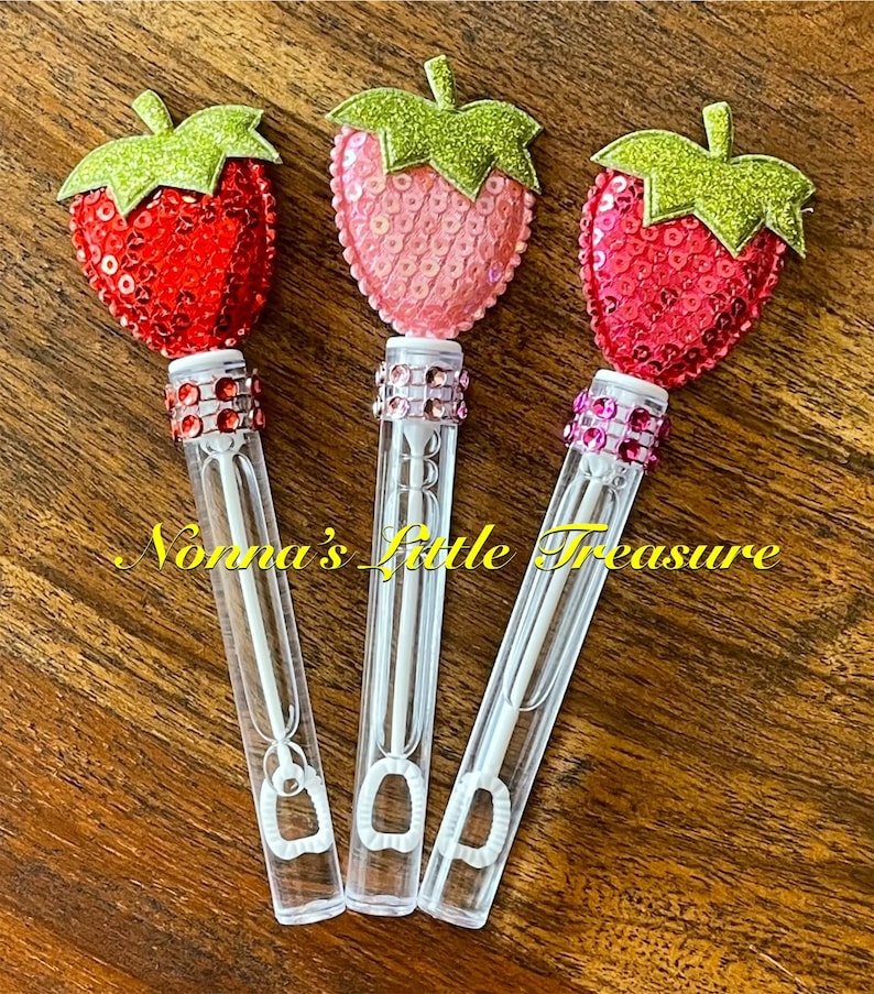 May include: Three bubble wands with strawberry-shaped tops. The tops are made of red, pink, and fuchsia sequins with green glitter leaves. The wands are clear plastic with a white wand inside.