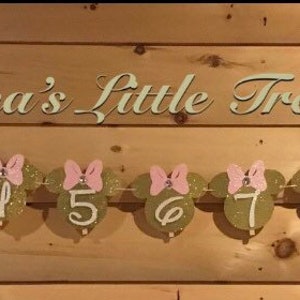 May include: A gold glitter Minnie Mouse themed banner with pink bows and white numbers 1 to 12. The banner reads "Nonna's Little Treasure".