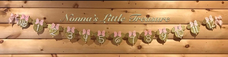 May include: A gold glitter Minnie Mouse themed banner with pink bows and white numbers 1 to 12. The banner reads "Nonna's Little Treasure".